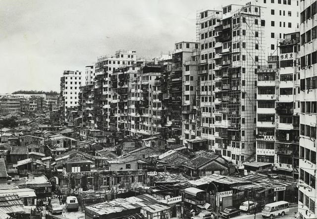 Black-and-white photo of tall, crowded apartment blocks rising behind low rooftop shacks, 1975.